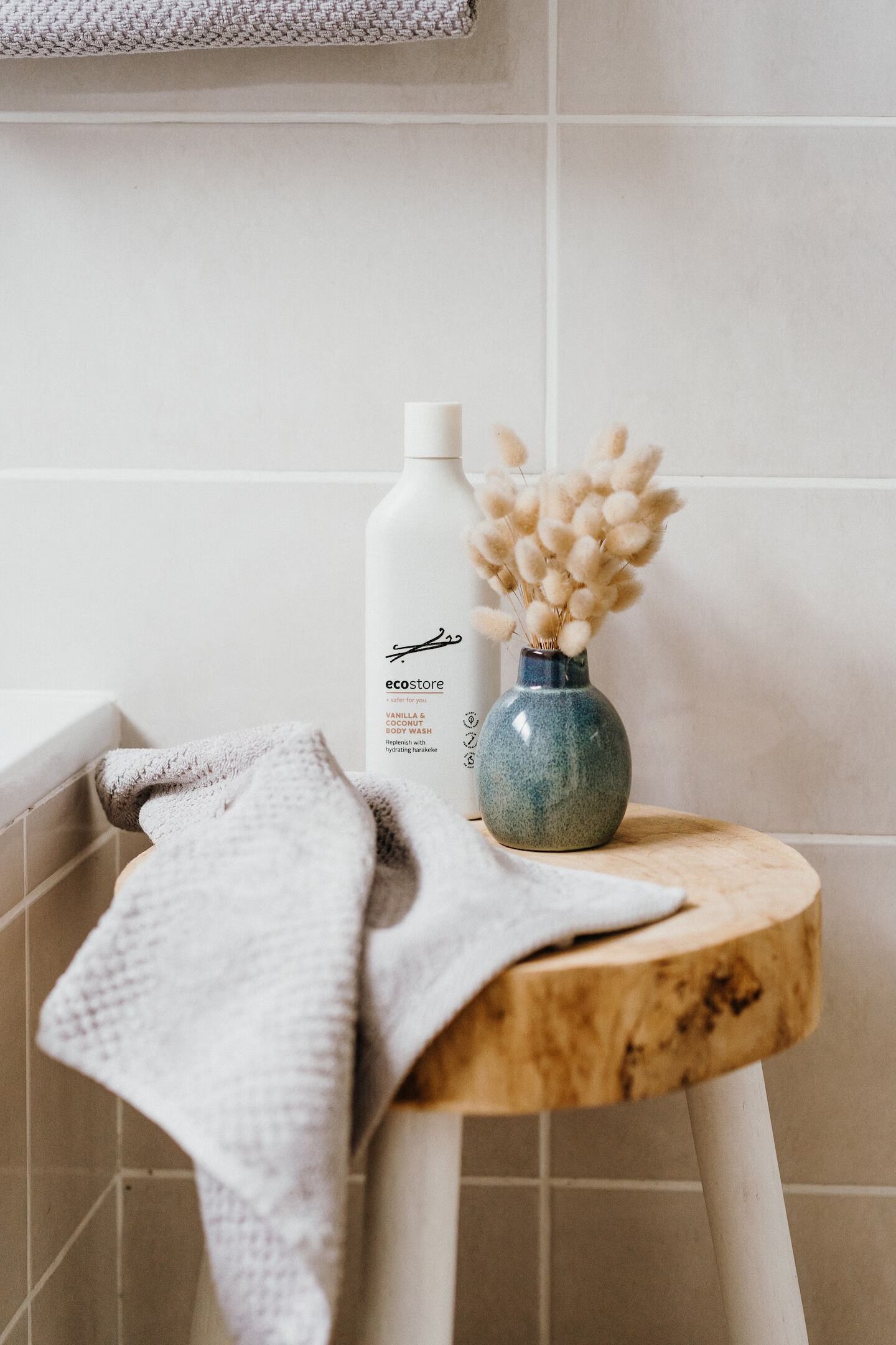 A bathroom with a white bathtub, a wooden stool, a blue vase with dried pampas grass, and a bottle of body wash.