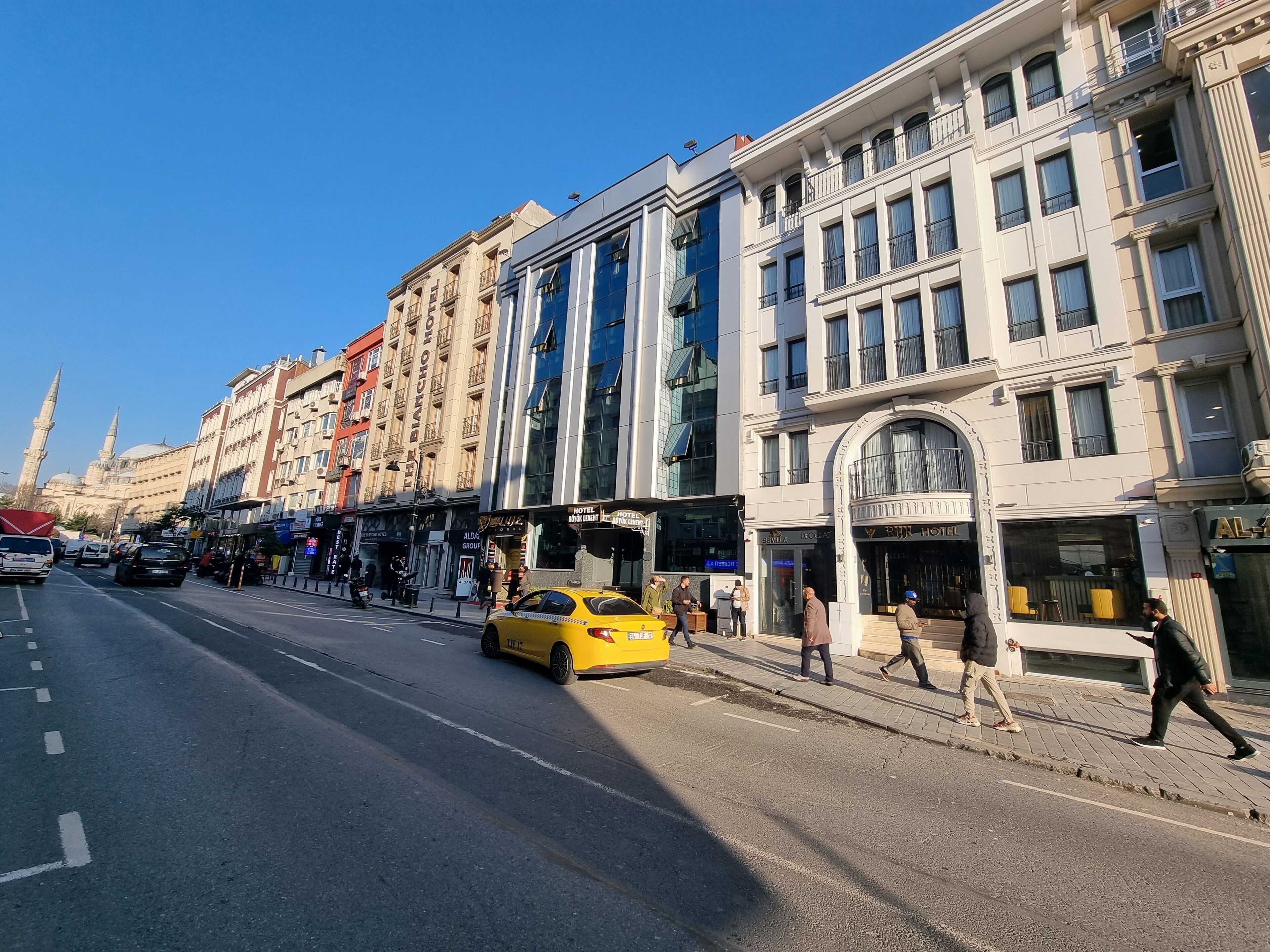A street with a yellow taxi, buildings with shops, and a mosque in the background.