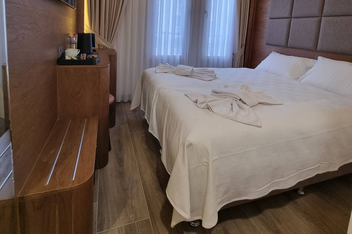 A hotel room with a bed, a TV mounted on the wall, a wooden bench, and a window with curtains.