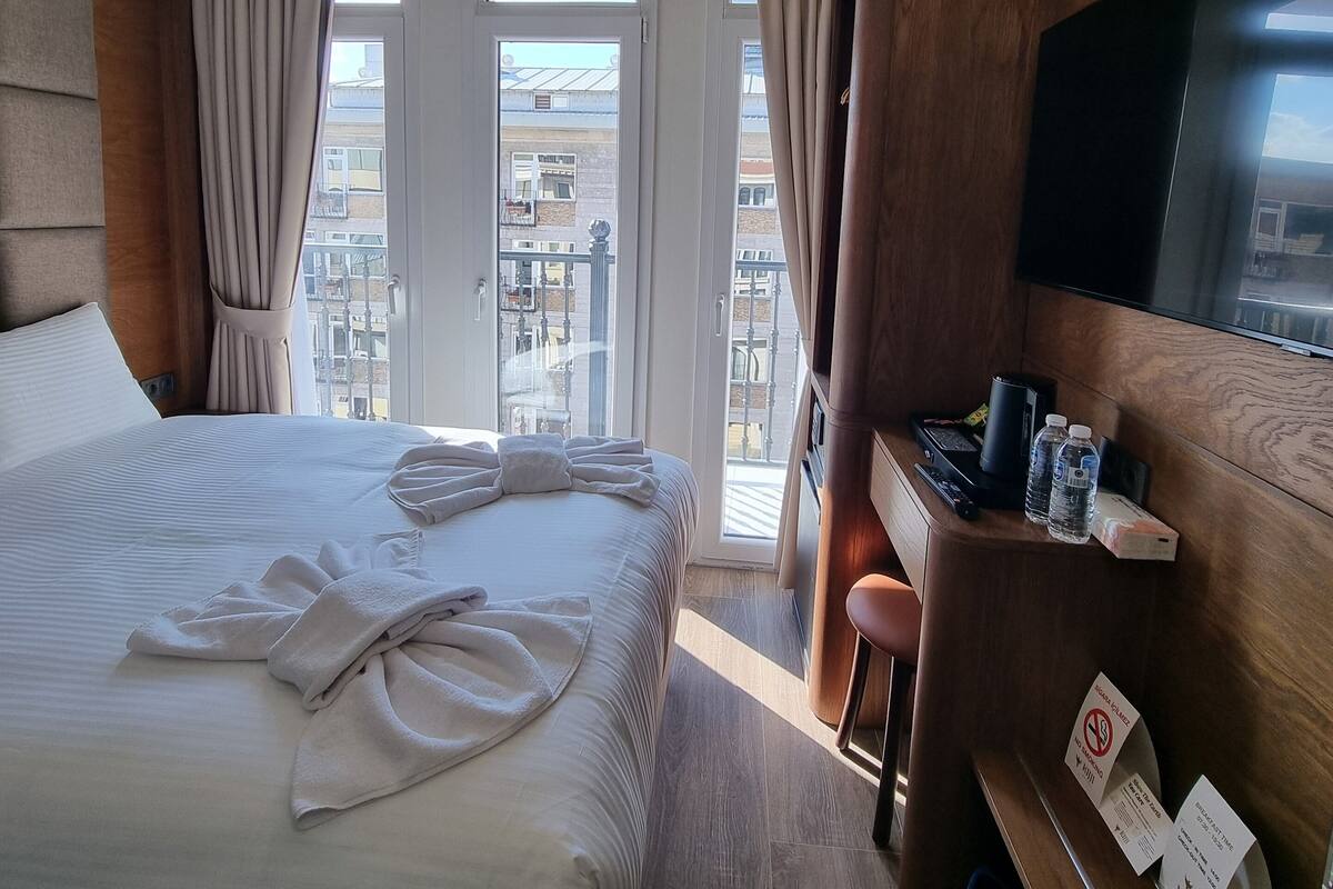 A hotel room with a large bed, a desk with a flat-screen TV, and a balcony with a view of the city.
