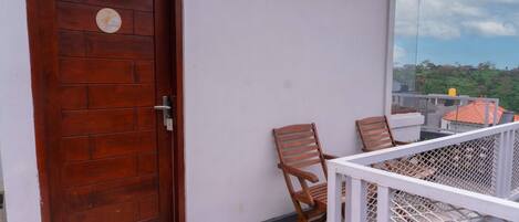 A wooden door with a circular emblem, a white wall, and a balcony with a white railing and two wooden chairs.