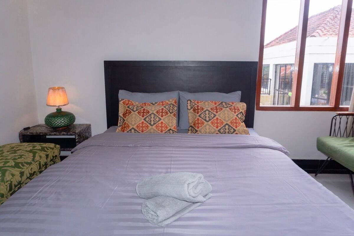 A neatly made bed with patterned pillows, a bedside lamp, and a view of the outside through a window.