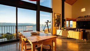 Un amplio espacio habitable con un gran ventanal que da a una masa de agua, una mesa de comedor de madera y una estantería llena de libros.