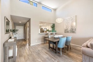 Un vaste espace dédié à la salle à manger, avec une table et des chaises en bois, une cuisine à l’arrière-plan et une grande fenêtre qui laisse entrer la lumière naturelle.