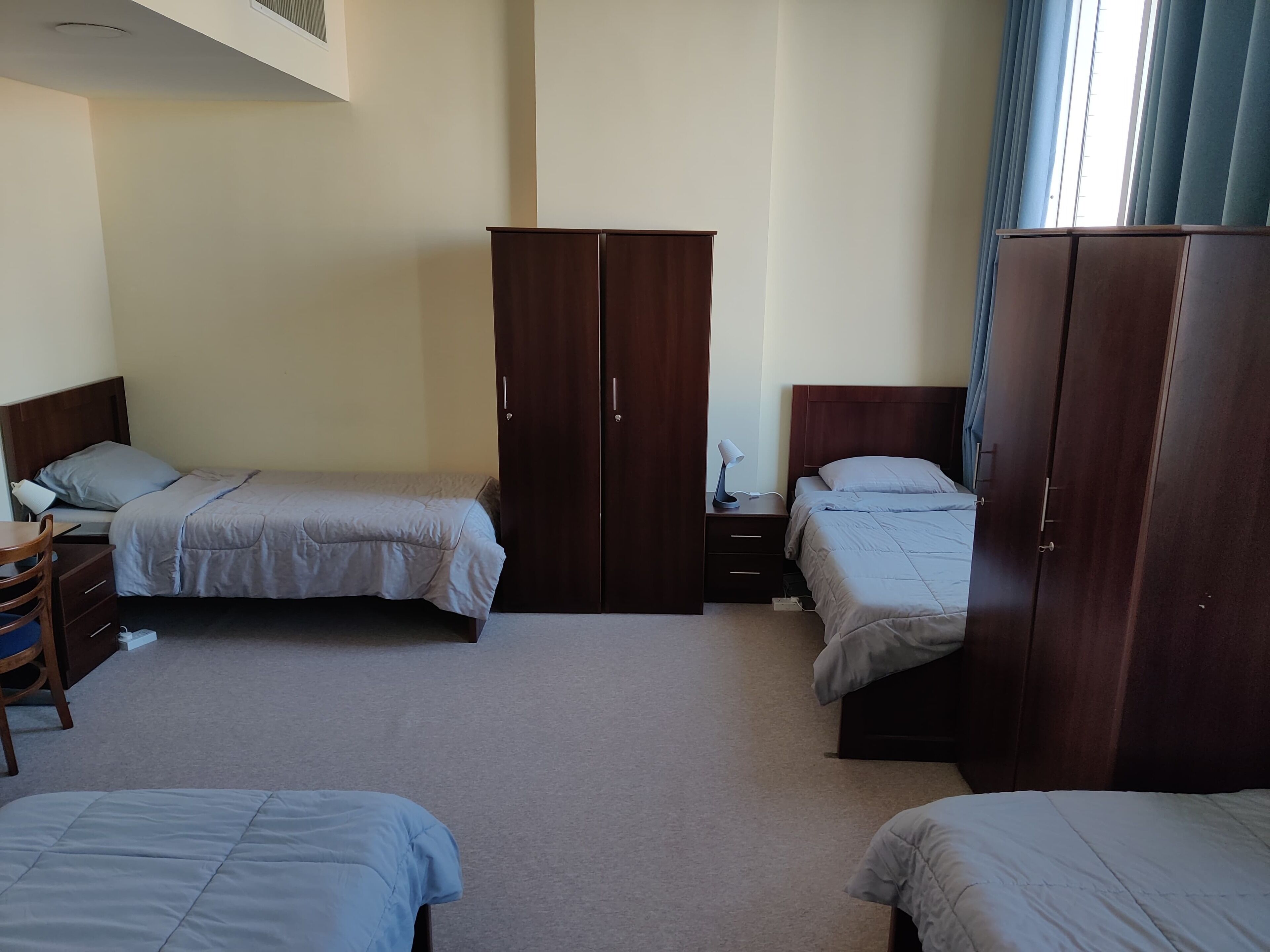 A hotel room with two single beds, a wooden wardrobe, a desk with a chair, and a nightstand with a lamp.