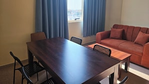 A room with a dining table, chairs, a sofa, and a window with curtains. - Oasis Hostel (Abu Dhabi)