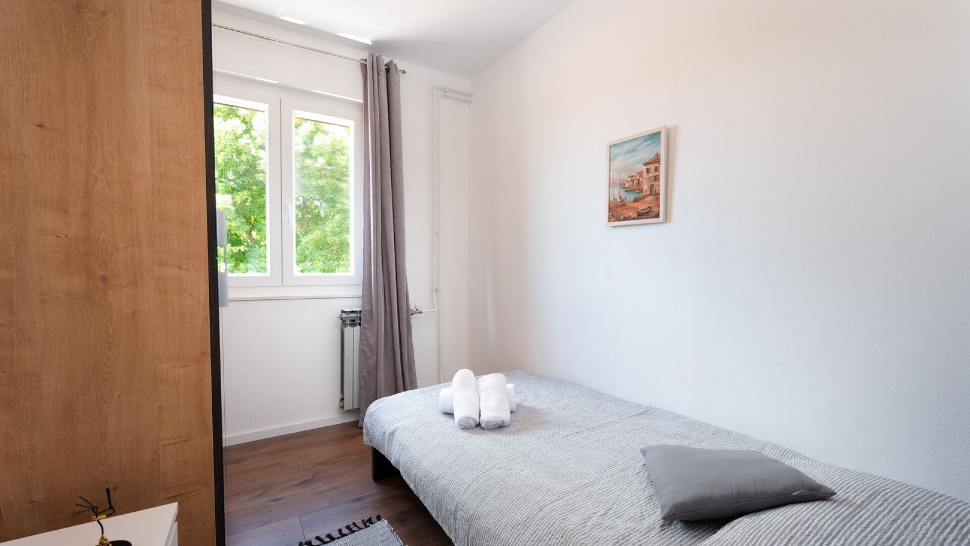 Ein Schlafzimmer mit einem Bett, einem Heizkörper, einem Fenster mit Vorhängen, einem Gemälde an der Wand und einem Holzboden.