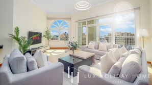 A modern living room with a flat-screen TV, a coffee table, and a sofa. There is a large window with a view of buildings. - LUX The Shoreline Palm Island Suite 2 (Dubai)