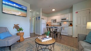 A living room with a sofa, a chair, a round table with a book and a plant, a kitchen area with cabinets and appliances, and a TV on the wall.
