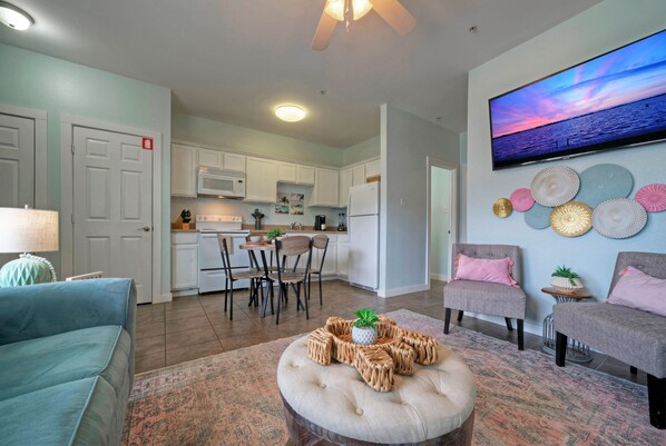 A modern living room with a sofa, ottoman, and chairs, a kitchen in the background, and a TV mounted on the wall.