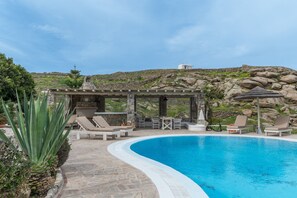 A pool with lounge chairs, a pergola, and a view of rocky hills. - Apartment 'Myconian V' with Sea View, Wi-Fi and Air Conditioning (Ornos)