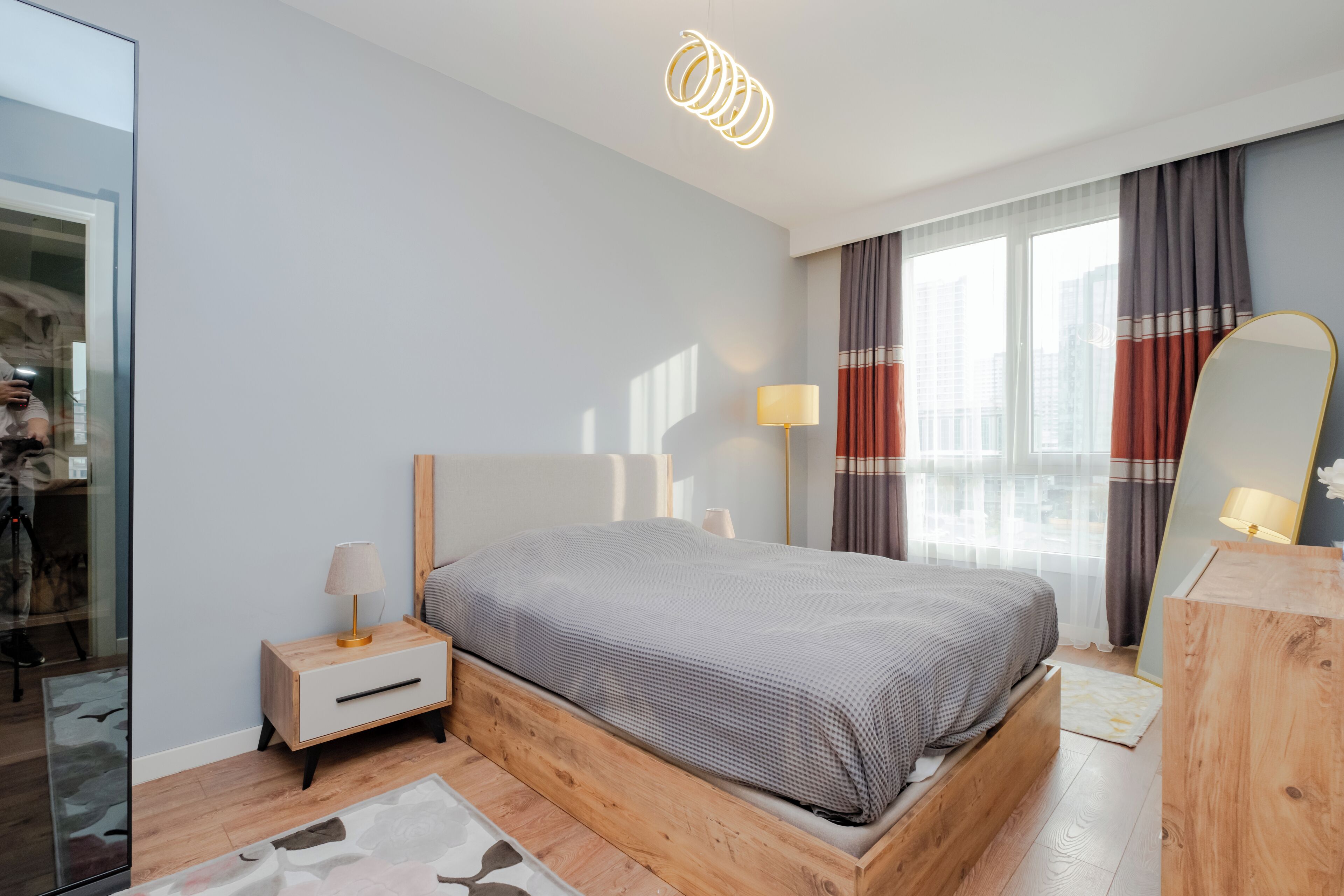 Ein modernes Schlafzimmer mit einem Holzbett, Nachttischen, einem Spiegel und Blick auf die Stadt durch große Fenster.