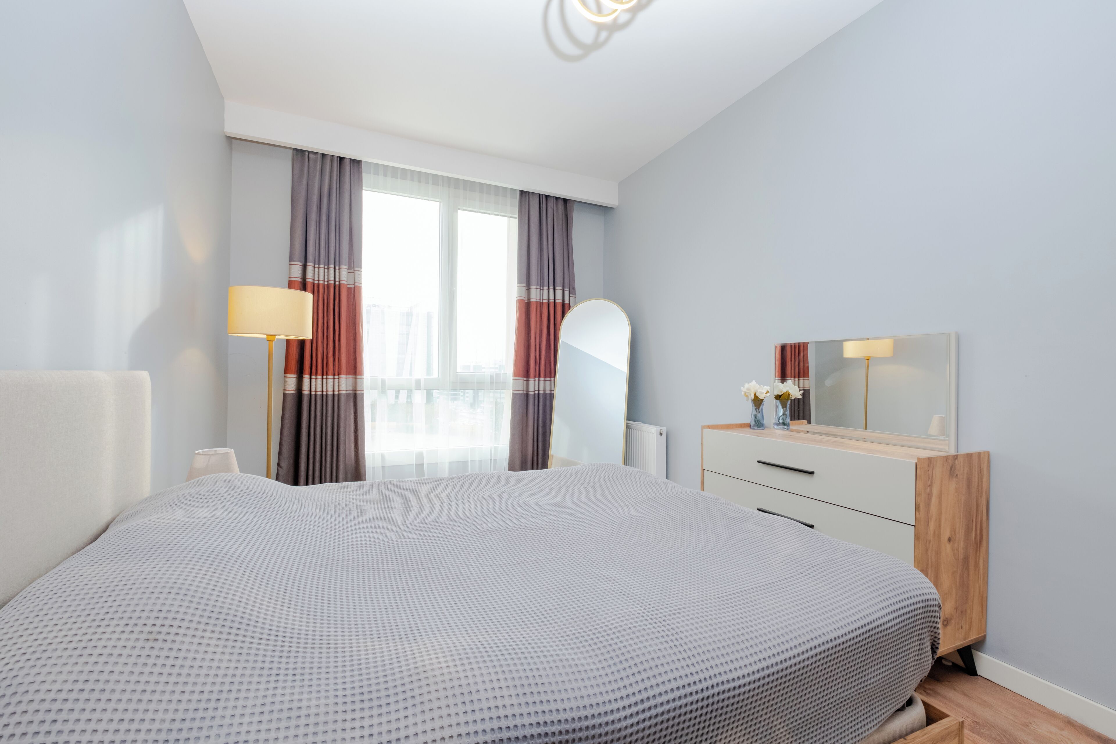 Une chambre moderne avec un grand lit, une commode en bois, un miroir et une fenêtre avec des rideaux.