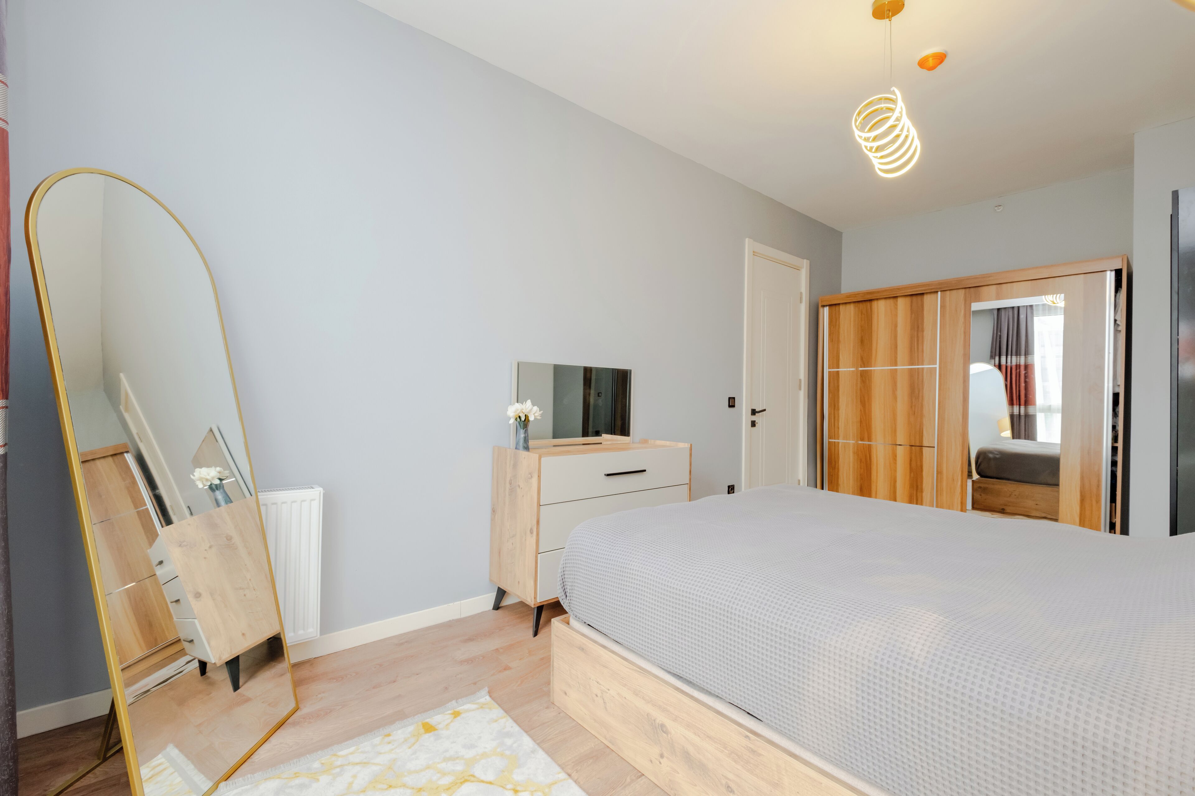 A modern bedroom with a large bed, a wooden dresser, a mirror, and a wardrobe.