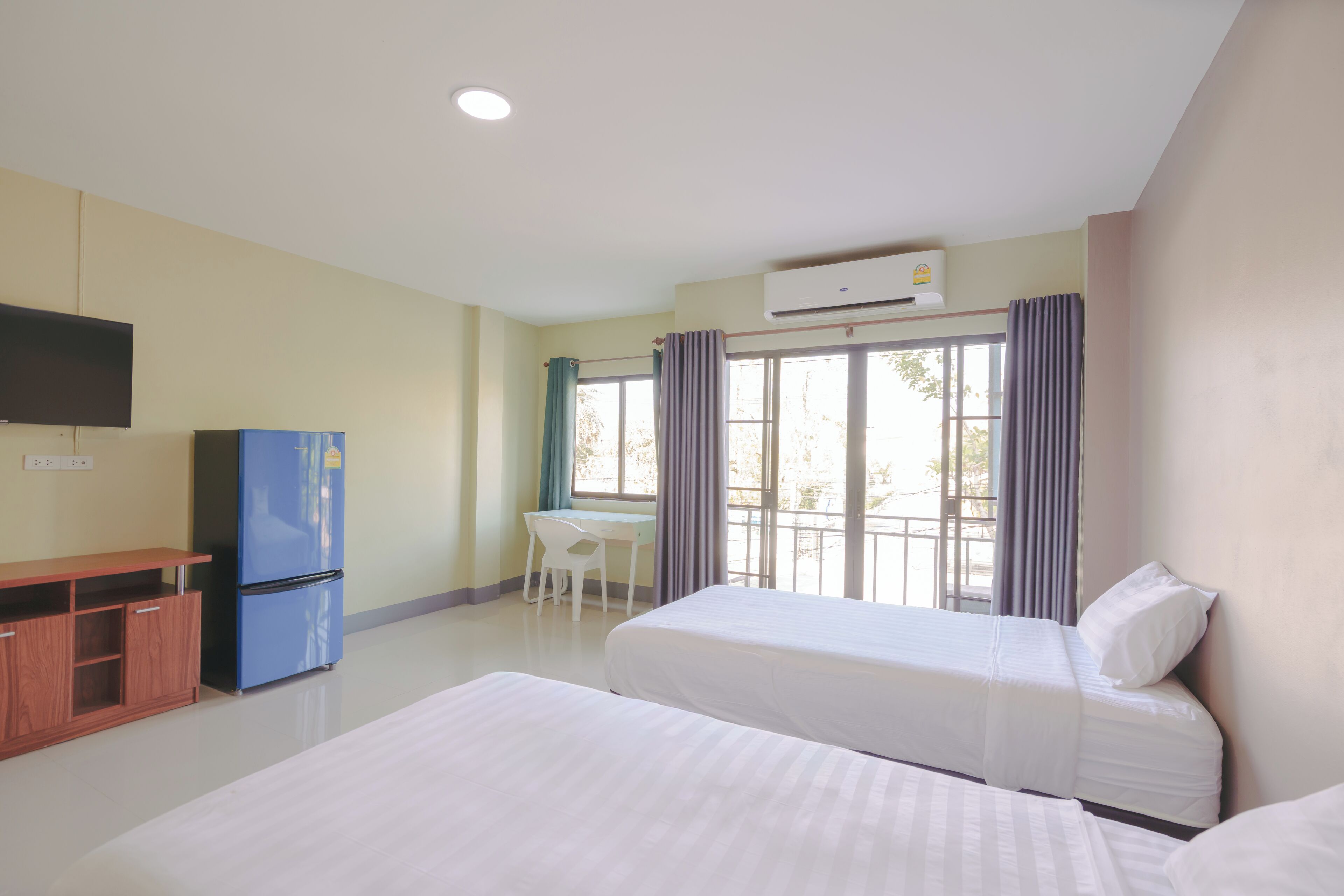 A hotel room with two beds, a desk, a blue refrigerator, and an air conditioning unit.
