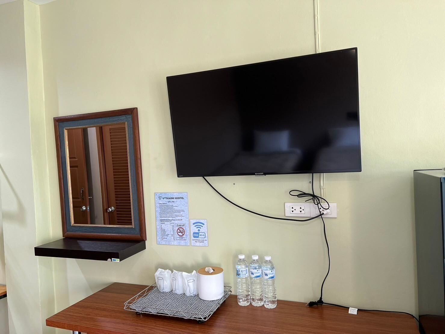 A room with a mounted flat-screen TV, a wooden door, a small table with toiletries, and a water bottle.