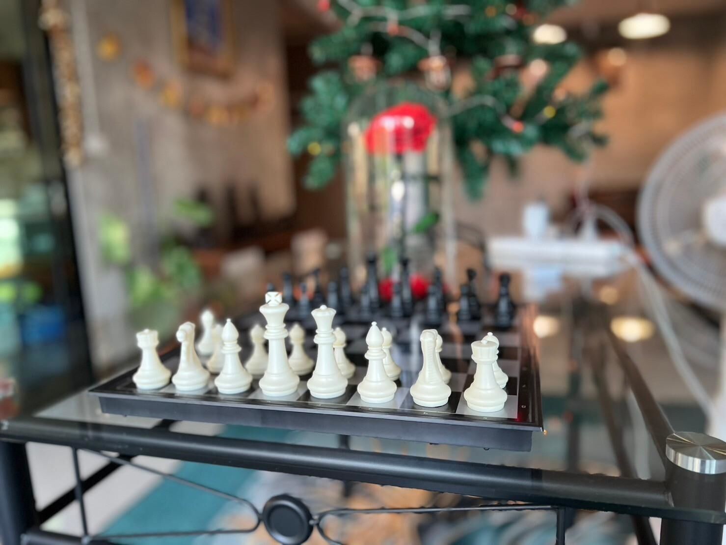A chessboard on a glass table with a blurred background including a plant and a fan.