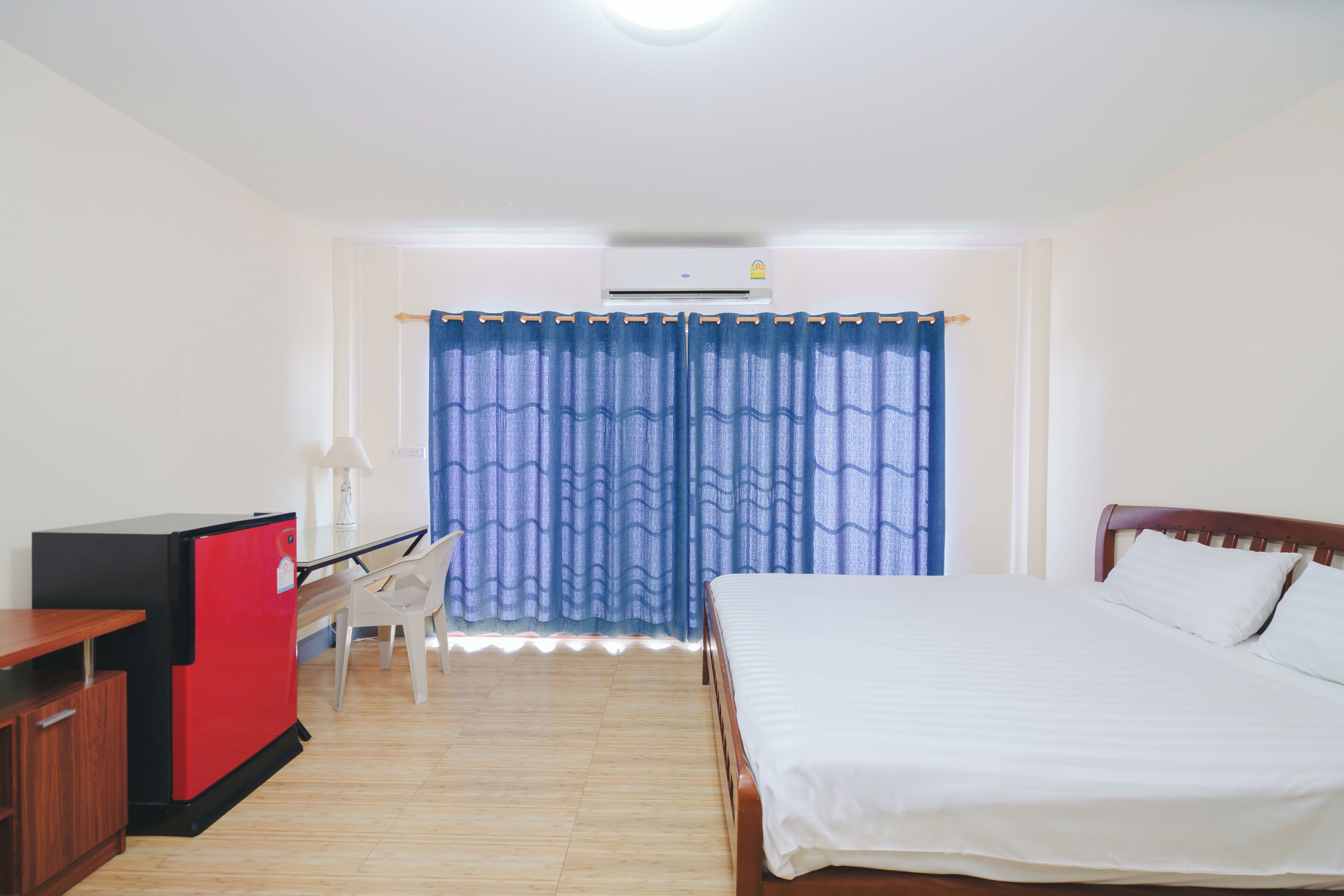 A hotel room with a bed, a desk with a red refrigerator, a chair, and a window with blue curtains.