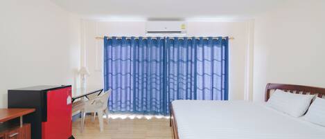 A hotel room with a bed, a desk with a red refrigerator, a chair, and a window with blue curtains.
