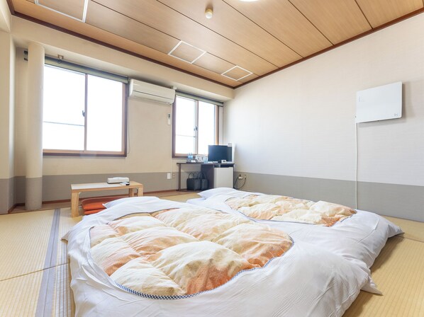 A hotel room with tatami flooring, a large bed with a patterned blanket, a desk with a chair, a television, and a window with curtains. - Hotel Boujosou (Kazuno)