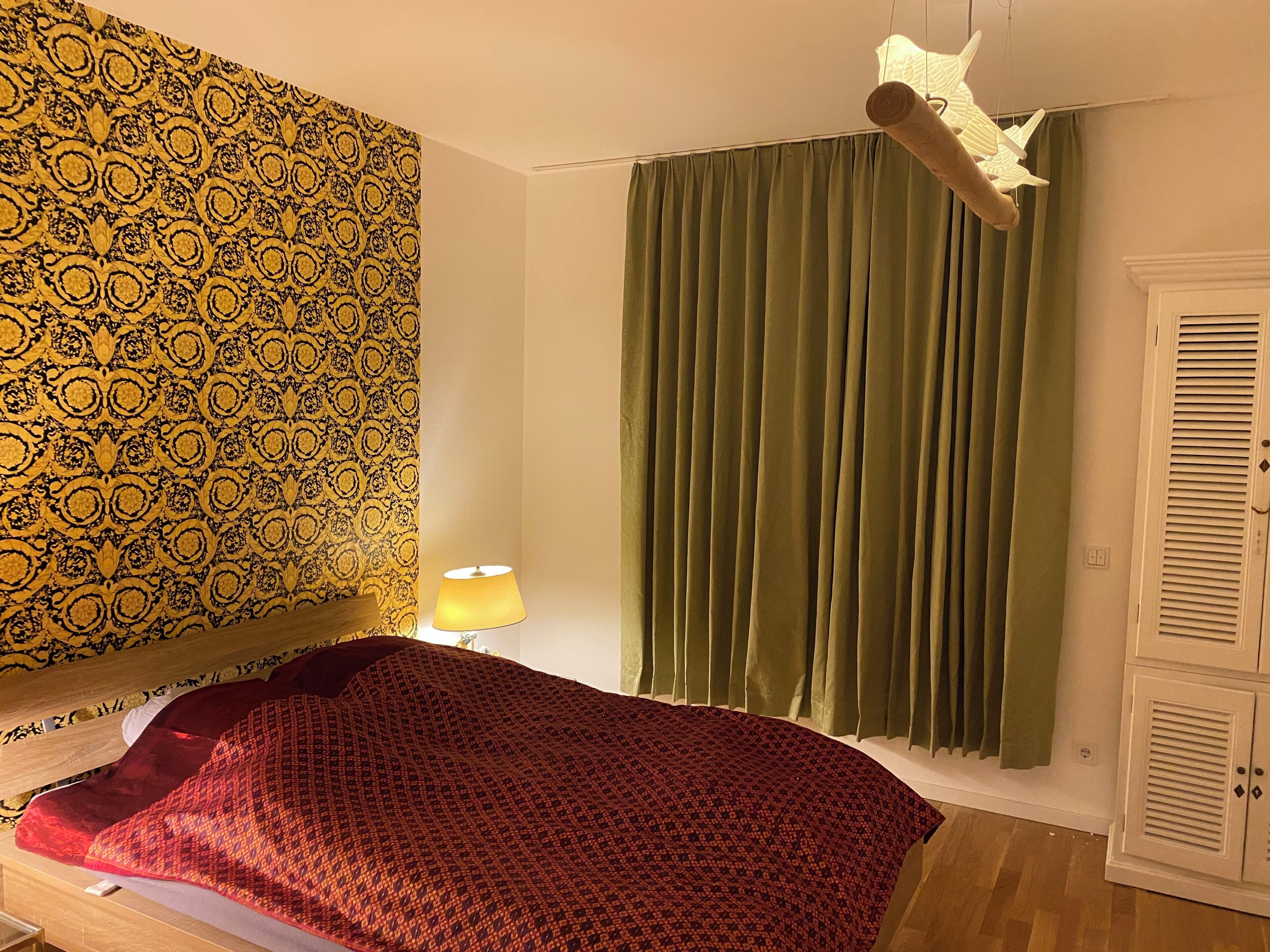 Une pièce avec un papier peint à motifs, un lit recouvert d’une couverture rouge, un rideau vert et une armoire blanche.