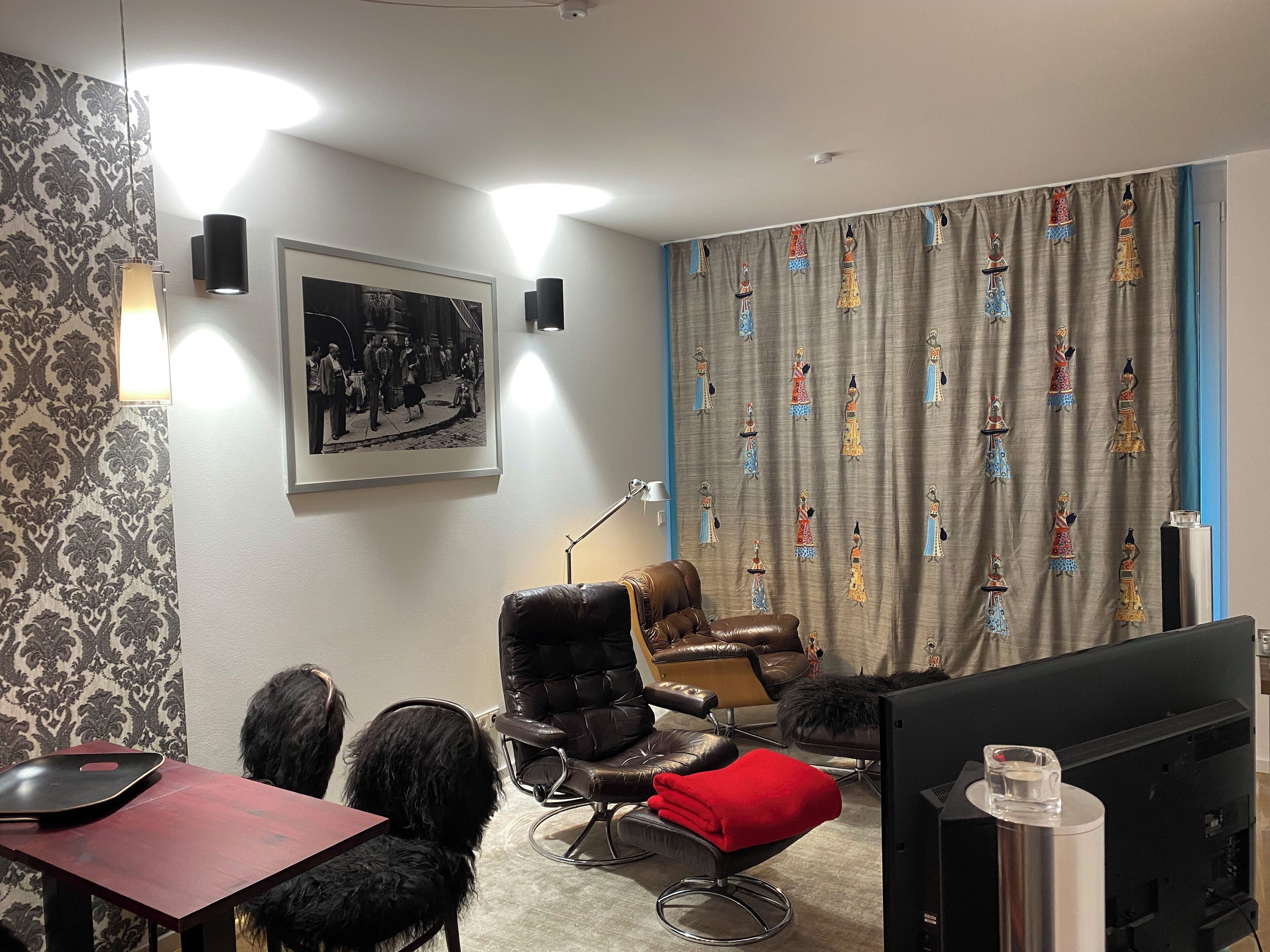 Une pièce avec un fauteuil en cuir marron, un coussin rouge, une photographie en noir et blanc encadrée et un rideau aux motifs colorés.