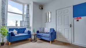 Una habitación con dos sofás azules, una mesita, una planta en maceta y una puerta con el letrero 'No Smoking'.