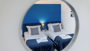 Habitación de hotel con dos camas, cada una con cabecero azul y ropa de cama blanca. Entre las camas hay una mesita de noche con una lámpara.