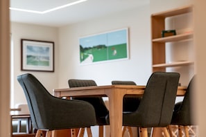 Un coin repas avec une table en bois et quatre chaises rembourrées. Des tableaux encadrés ornent le mur, et une étagère expose divers objets.