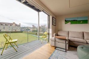 Una sala de estar moderna con un gran ventanal corredera que da a un balcón, un sofá color beige, una mesa de centro de madera y un cuadro con un paisaje enmarcado.