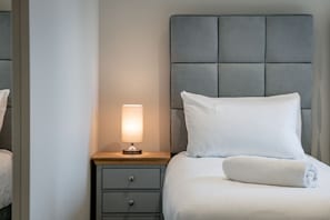 A neatly made bed with a white pillow and a rolled white towel, a bedside lamp, and a gray headboard.