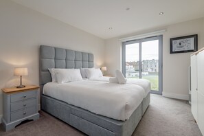 A modern bedroom with a grey upholstered headboard, white bedding, a bedside table with a lamp, and a large window offering a city view.