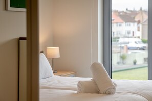 A hotel room with a neatly made bed, a bedside lamp, and a view of the street through a large window.