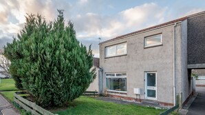 Un edificio residencial de dos pisos con un gran árbol siempreverde en el jardín delantero, una ventana pequeña y una puerta con el número 70.
