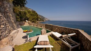 A stone pool with lounge chairs and a table, overlooking a coastal view.