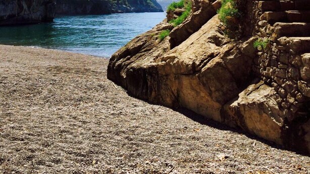 A coastal view with a rocky cliff, a clear blue sea, and a mountainous backdrop.