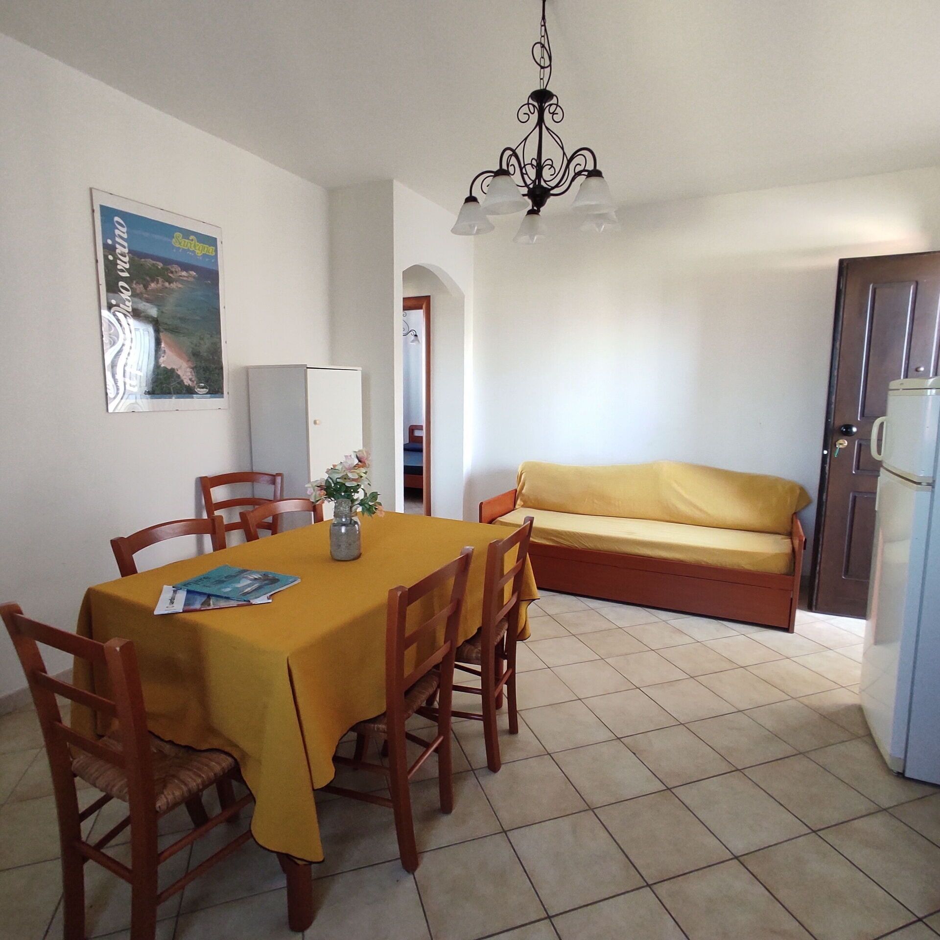 A dining area with a table set for four, a sofa, a refrigerator, and a chandelier.