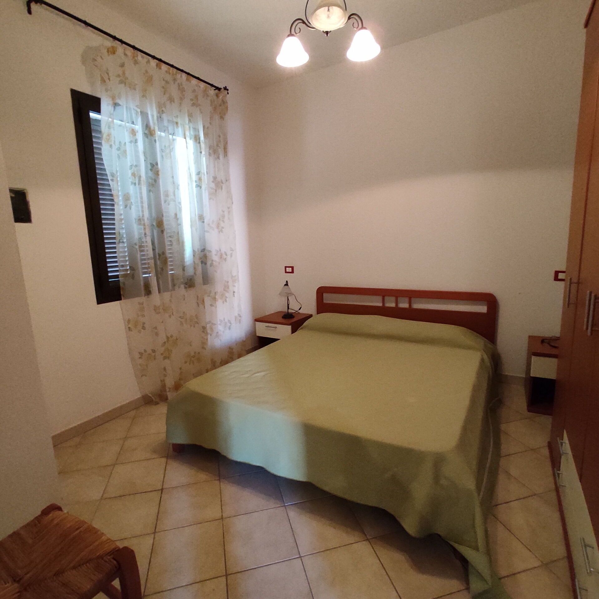 Une chambre à coucher comprenant un lit, une table de chevet, une lampe, une fenêtre avec des rideaux et une armoire en bois.