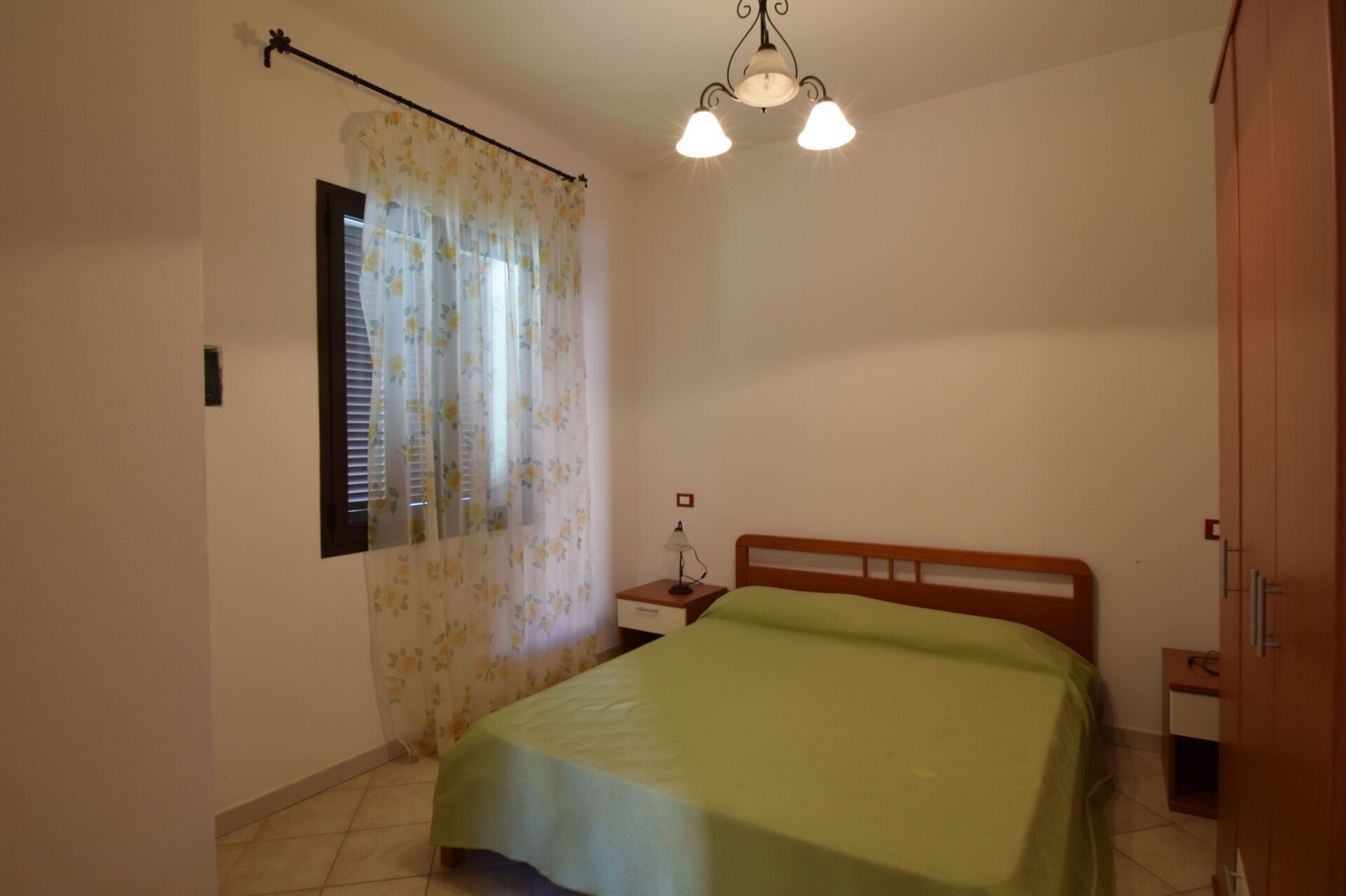 Une chambre à coucher comprenant un lit, une table de chevet, une lampe, une fenêtre avec des rideaux à motifs floraux et un luminaire au plafond.