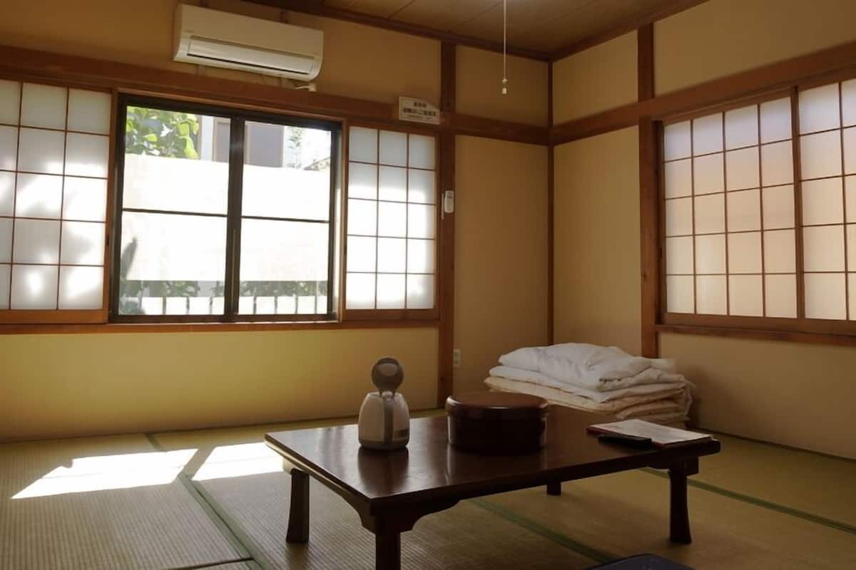 Une pièce de style japonais traditionnel, avec un sol recouvert de tatamis, une petite table sur laquelle se trouvent une bouilloire et un livre, et une fenêtre dotée de portes coulissantes en papier de riz (shoji).