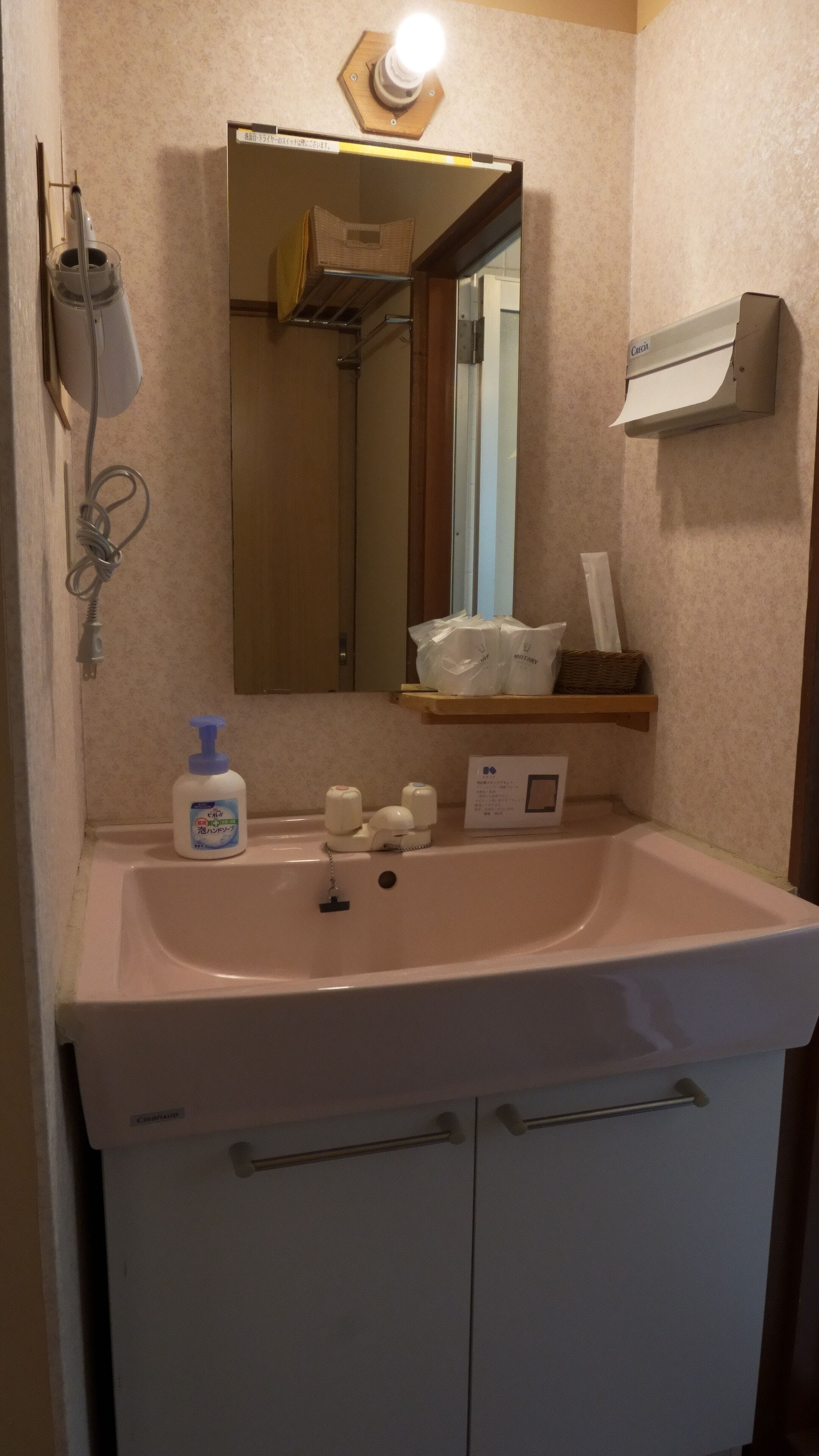 Een badkamer met een wastafel, spiegel, zeepdispenser, handdroger en een plank met toiletartikelen.