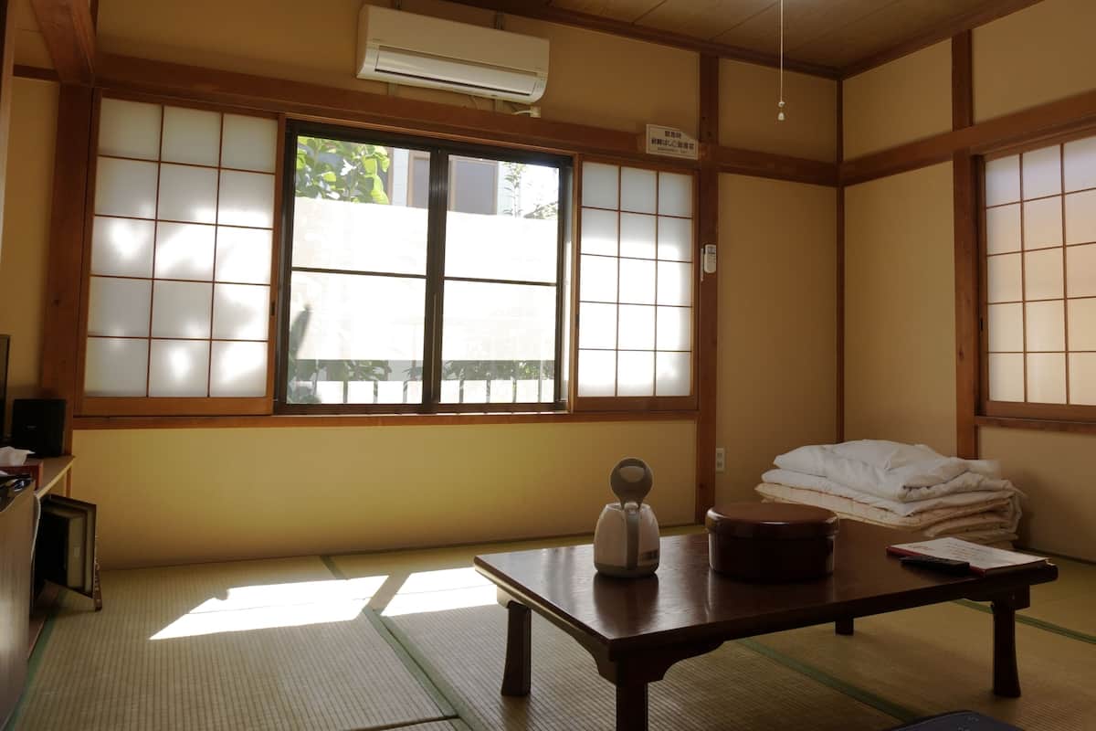 Une pièce de style japonais traditionnel, avec un sol recouvert de tatamis, un paravent en papier de riz (shoji), un futon, une table avec une bouilloire et un radiateur.