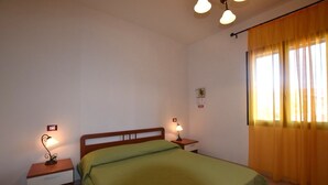 A single bed with a green bedspread in a room with two bedside tables, each with a lamp. There is a window with yellow curtains on one side. - Agnese Residence Sardinia - Trilo 2 (Valledoria)