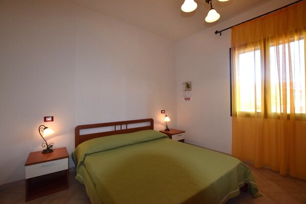 Un lit simple avec une couvre-lit verte dans une chambre où se trouvent deux tables de chevet, chacune étant surmontée d’une lampe. Une fenêtre avec des rideaux jaunes se trouve sur un des murs.