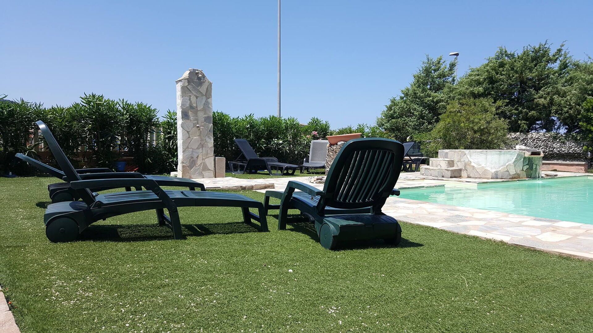 Un espace aménagé au bord de la piscine, avec des chaises longues, un pilier en pierre et un ciel d’un bleu limpide.