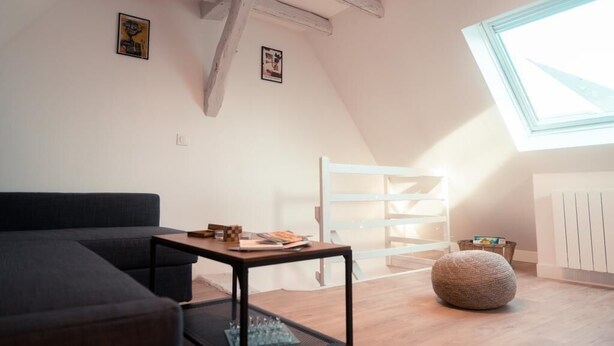 Un dormitorio estilo loft moderno con techo inclinado, un tragaluz grande, un sofá oscuro, una pequeña mesa con libros, una silla blanca y un otoman redondo.