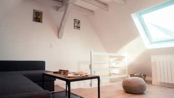 Un dormitorio estilo loft moderno con techo inclinado, un tragaluz grande, un sofá oscuro, una pequeña mesa con libros, una silla blanca y un otoman redondo.