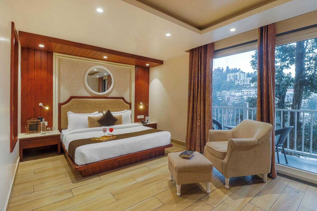 A hotel room with a large bed, a round mirror, a nightstand, a chair, and a balcony with a view of buildings.