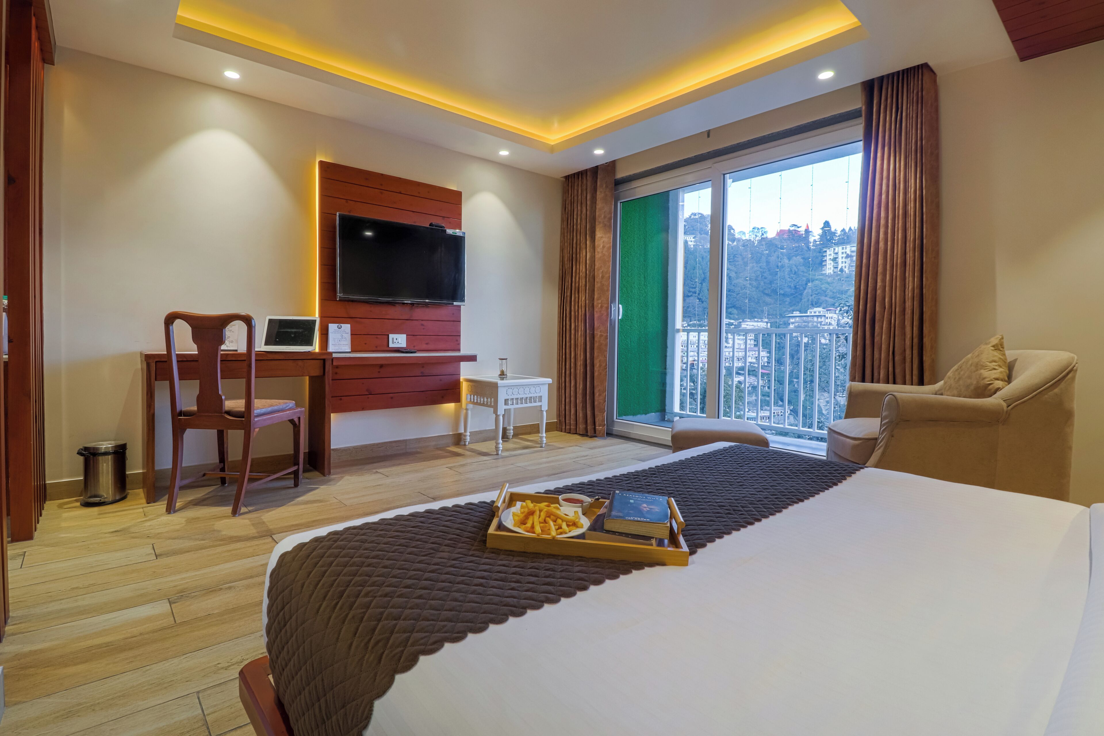 A hotel room with a bed, a desk with a laptop, a TV, and a view of a cityscape through a large window.