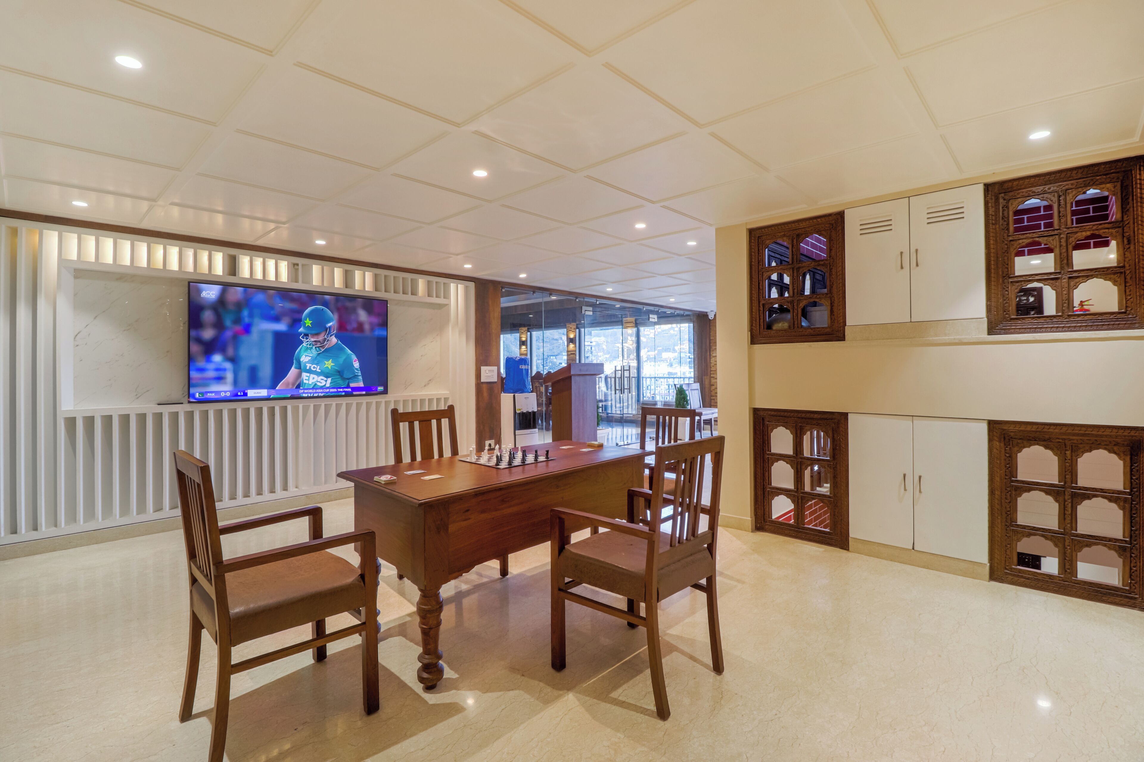 A dining area with a wooden table and chairs, a television mounted on the wall, and a view of the city through large windows.